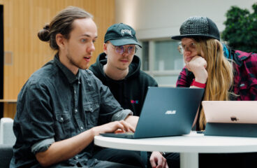 Kolme opiskelijaa kokoontuneena pöydän ääreen ja työskentelemässä kannettavilla tietokoneilla.
