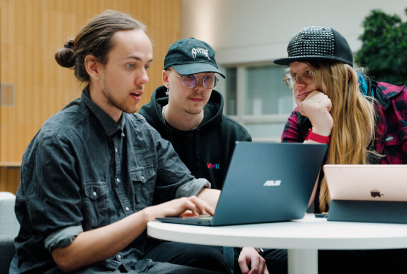 Kolme opiskelijaa kokoontuneena pöydän ääreen ja työskentelemässä kannettavilla tietokoneilla.